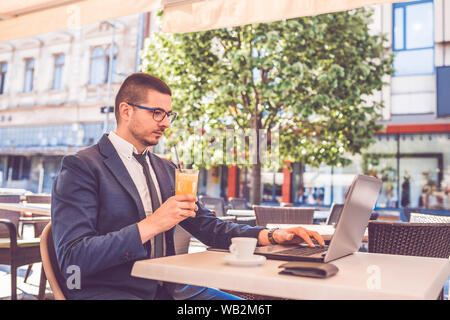 Young professional businessman wearing suit, drinking fresh orange juice and using  laptop outdoors. Successful manager working in cafe during break a Stock Photo