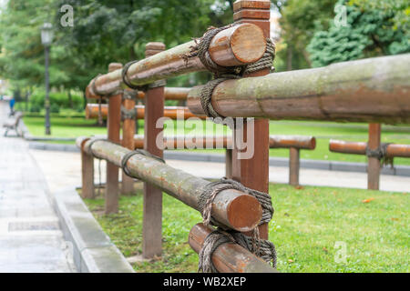 A wooden log fence fixed with a rope in a park next to an asphalt path with a concrete curb. Against the background of trees in summer Stock Photo