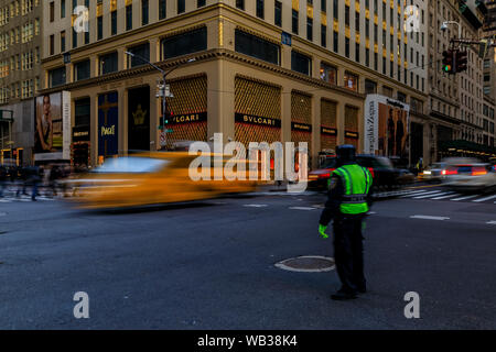 New York USA - Yellow taxi cabs and other blurred cars speed past people crowds and a traffic policeman, at a busy crossing on 5th Avenue in Manhattan Stock Photo