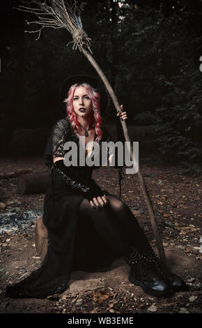 Halloween theme: contemporary young witch in black dress and hat with broom sitting in a dark grove Stock Photo