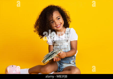 Cute afro girl holding bunch of money, yellow background Stock Photo