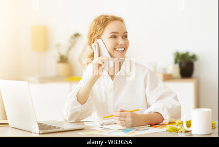 Happy seamstress talking on phone, discussing outfit with customer Stock Photo