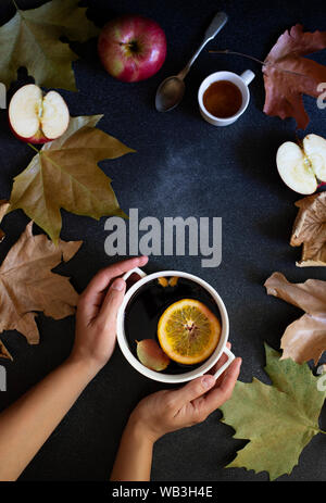 Female hands holding mug with mulled wine. Autumn and winter warm alcoholic drink. Autumn leaves, honey, apples, oranges, vintage dishes. Autumn mood Stock Photo