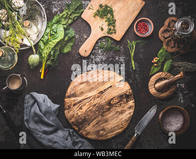 Vegetarian food cooking background with chard leaves , spices and kitchen utensils around empty circle wooden cutting board on dark rustic background. Stock Photo