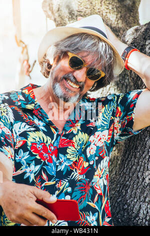 Mature man smiling and enjoying taking selfies with his mobile phone, wearing a vintage hat next to a tree Stock Photo