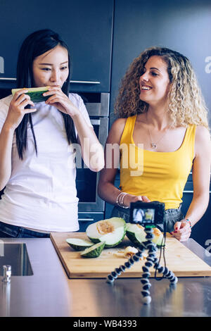 Asian young girl tasting a piece of melon, another smiling young woman is cutting the melon while they are recording with a camera Stock Photo