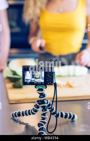 Young blonde girl cutting a melon with a knife in the kitchen, while recording with a camera. Selective focus Stock Photo
