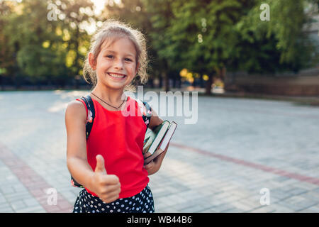 Happy pupil girl wearing backpack and holding books. Smiling kid showing thumb up outdoors primary school. Education Stock Photo