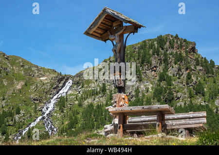 Crucifix, wooden Christ sculpture, wooden bench in Leibnitztal near Hochschober Hut. Austrian Alps. Stock Photo