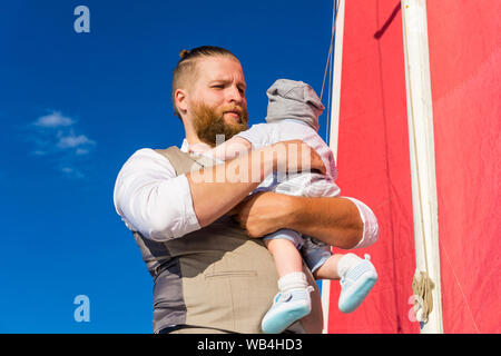 bearded man holding a baby in his arms against the background of blue sky and a ship mast with red sails Stock Photo