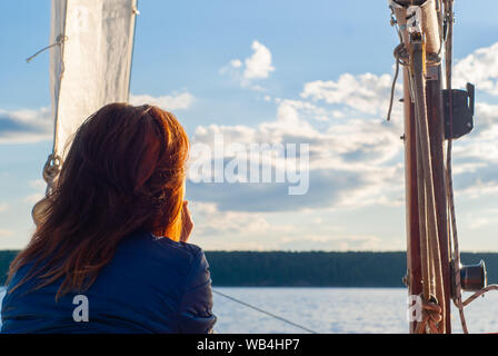 red-haired woman on a sailboat admiring the sunset sky and distant wooded shore