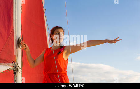 woman in red dress with anxious facial expression standing on a sailboat points hand forward Stock Photo