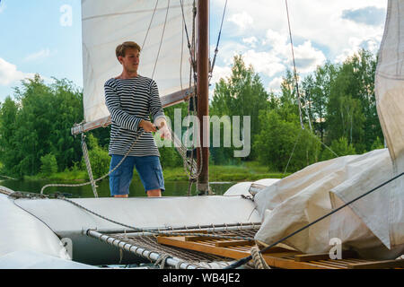 young sailor raises a sail on a small sailing catamaran moored in a wooded bay Stock Photo