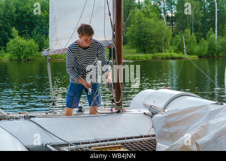 young sailor raises a sail on a small sailing catamaran moored in a wooded bay Stock Photo