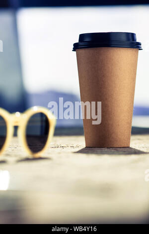 Disposable coffee cup on the rough table next to sunglasses. Sunny summer morning and holiday mood. Stock Photo