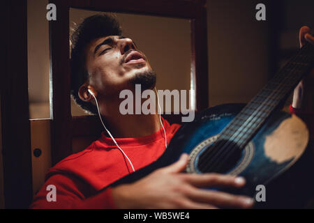 Young handsome man having fun listening to music and playing guitar with phone headset in room. Stock Photo