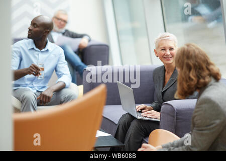 Portrait of successful senior businesswoman smiling cheerfully while talking to colleagues and using laptop during meeting in office hall, copy space Stock Photo
