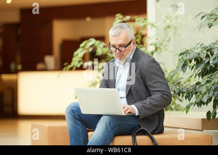 Portrait of contemporary senior businessman using laptop while working in lobby or office hall, copy space Stock Photo