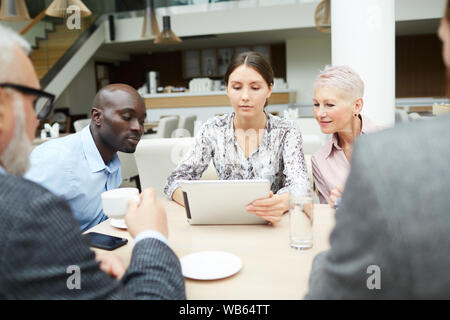 Diverse group of business people discussing work during meeting, focus on young woman using digital tablet sitting at table Stock Photo