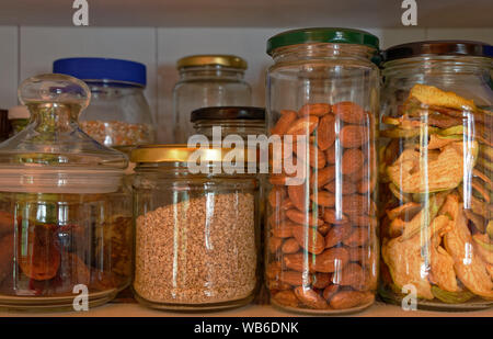 Glass storage jars lined up in a cupboard filled with nuts, dried apples and grains Stock Photo