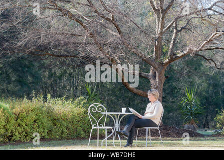 A middle aged white woman sits at a white outdoor table and chairs in morning winter sunlight in a garden with a cup of tea reading a book Stock Photo