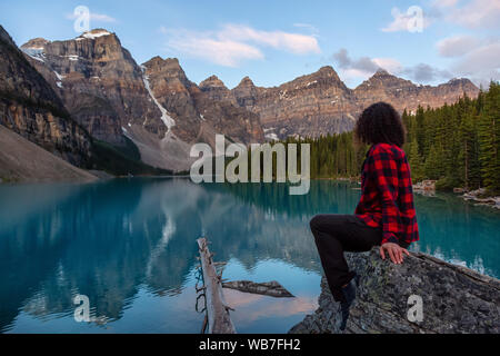 Girl enjoying the beautiful view of an Iconic Famous Place, Moraine Lake, during a vibrant summer sunrise. Located in Banff National Park, Alberta, Ca Stock Photo