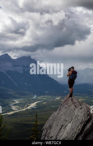 Adventurous Girl taking pictures on top of a rocky mountain during a cloudy and rainy day. Taken from Mt Lady MacDonald, Canmore, Alberta, Canada. Stock Photo