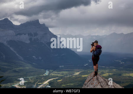Adventurous Girl taking pictures on top of a rocky mountain during a cloudy and rainy day. Taken from Mt Lady MacDonald, Canmore, Alberta, Canada. Stock Photo