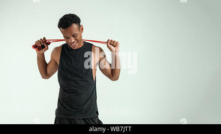 Healthy lifestyle. Strong and sporty young afro american man holding a jumping rope at his shoulders and smiling while standing against grey background. Sport equipment. Web banner Stock Photo