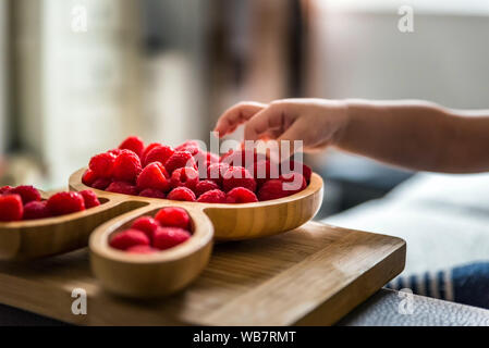 baby boy hands touch and take raw fresh raspberries on wooden bamboo plate indoor. baby exploring fruit