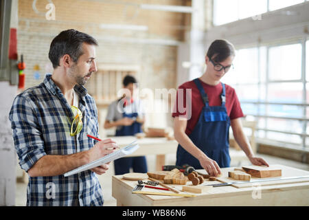 Waist up portrait of mature carpenter holding clipboard d and taking notes while overseeing trainees in workshop class, copy space Stock Photo