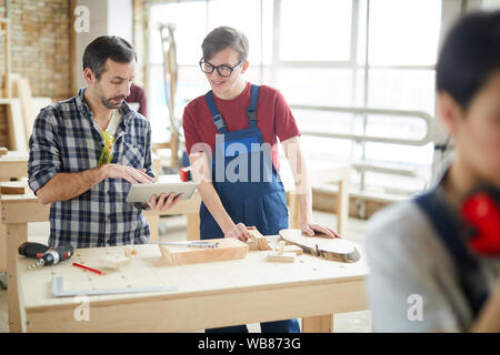 Waist up portrait of mature carpenter using digital tablet while talking to smiling young apprentice in workshop, copy space Stock Photo