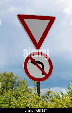 Give way and no left turn red and white signs mounted on strong metal pole surrounded with dense leaves on cloudy stormy blue sky background Stock Photo