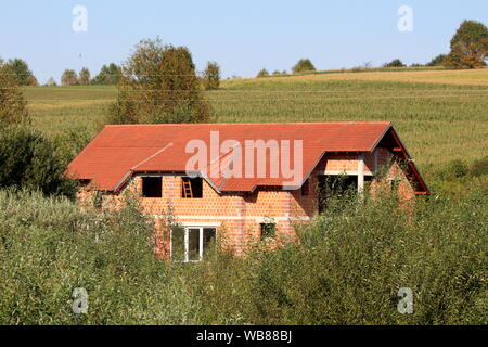 Large new unfinished red brick family house still under construction with new roof tiles and without windows completely surrounded with tall grass Stock Photo