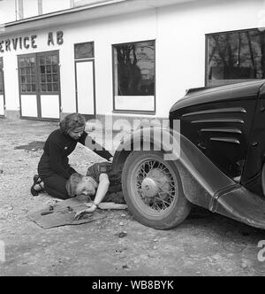 Service station of the 1950s. A young couple with their broken down car. He is trying to repair something in the front of the car and has his tools within range.  Sweden 1952. Kristoffersson ref BF75-8 Stock Photo