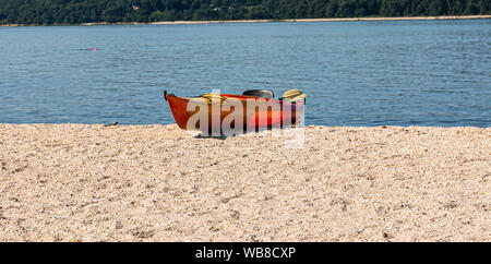 A single orange kayak on the sand of the beach with blue water in the background. Stock Photo