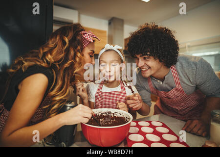 Happy father and his daughter are preparing cookies together in the kitchen. Father is pouring chocolate dough into molds and little girl wants to eat Stock Photo