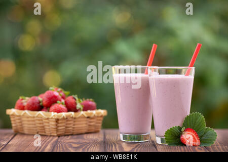 two glasses of strawberry milkshake or smoothie and fresh ripe berries in basket on wooden table outdoors Stock Photo