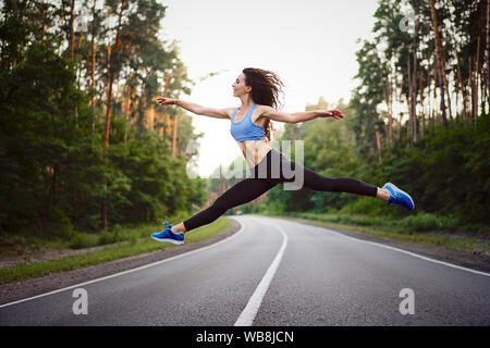 young beautiful brunette girl jumping in twine with background  asphalt road and pine forest Sportive woman showing happiness moments. Dancer training Stock Photo