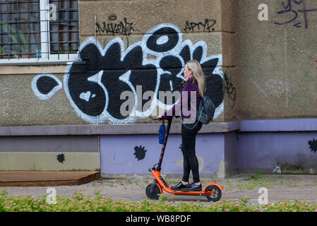 RIGA, LATVIA. 10th July 2019. Young woman with headphones riding with electric Fiqsy scooter on pavement in the city. Stock Photo