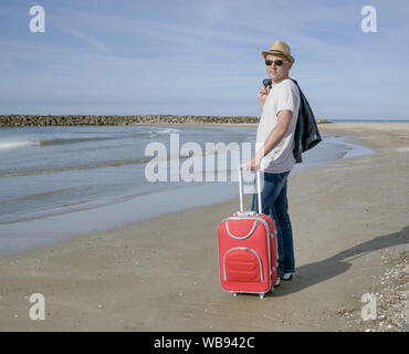 Young male tourist in a hat and with a red suitcase stands on the seashore, looking at the camera Stock Photo
