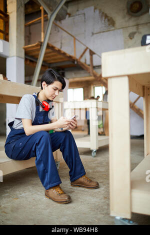 Full length portrait of young female worker using smartphone while resting on wooden stairs during break, copy space Stock Photo
