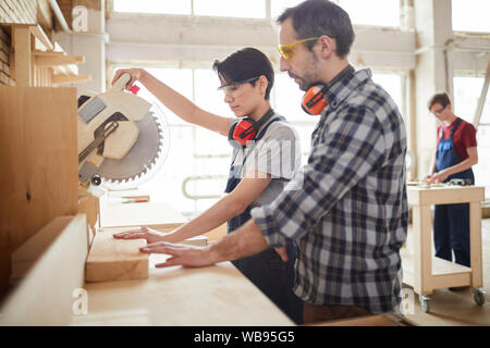 Side view portrait of mature carpenter helping young worker sawing wood in joinery workshop, copy space Stock Photo
