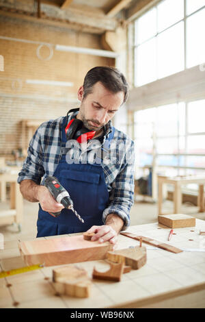 Waist up portrait of mature carpenter drilling wood while building handmade furniture in workshop Stock Photo