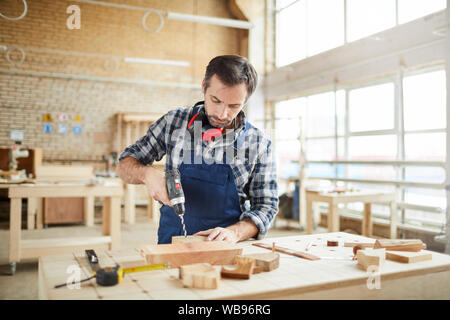 Waist up portrait of mature carpenter drilling wood while building handmade furniture in workshop, copy space Stock Photo