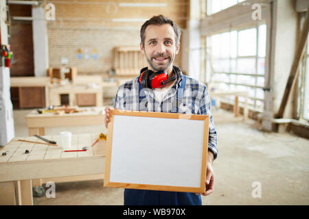 Waist up portrait of smiling mature carpenter holding blank frame and looking at camera, copy space Stock Photo