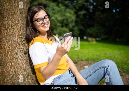 Lovely young girl resting against a park tree enjoys using smartphone Stock Photo