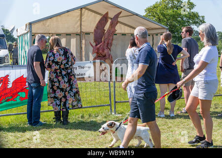Knutsford, Cheshire, UK. 25th Aug 2019. The 15th English Open Chainsaw Competition at the Cheshire County Showground, England - people walk past and view the winged creature being carved by Danny Thomas of Wales Credit: John Hopkins/Alamy Live News Stock Photo