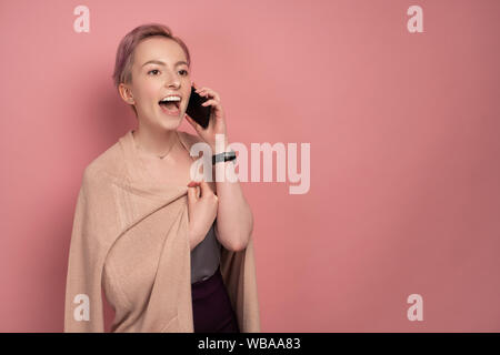 A girl with short pink hair in a cardigan joyfully opening her mouth, speaks on the phone, standing on a pink background. Stock Photo