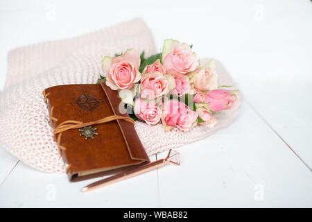 Pink roses laying and a leather notebook on a thick knit woolen cosy blanket laying on a white wooden background  Stock Photo